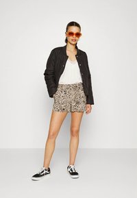 Casaco bomber preto acolchoado, blusa branca, calções bege com padrão, e ténis pretos. Modelo usa óculos de sol laranja, posando contra um fundo branco.