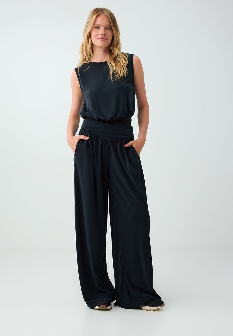 Femme aux longs cheveux blonds portant un haut noir sans manches et un pantalon noir à jambes larges, debout les mains dans les poches.