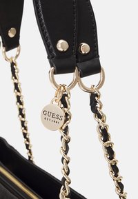 Black leather handbag with gold-tone chain and hardware. Features a tag with "GUESS EST.1981" and textured straps. Zipper closure visible.