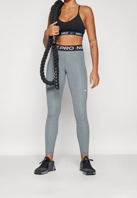 Szare legginsy z wysokim stanem o gładkiej fakturze i czarnym logo Nike, zestawione z czarnym stanikiem sportowym i czarnymi butami sportowymi.