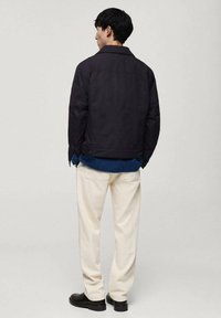 Marineblaues Jacket mit lässiger Passform, kombiniert mit einem blauen Hemd und cremefarbenen Hosen. Schwarze Schuhe runden das Outfit in neutralen Tönen ab.