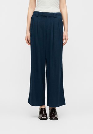 Kobieta ubrana w granatowe spodnie o szerokich nogawkach z plisami oraz jasnoszary, bezrękawnik, w zestawieniu z ciemnobrązowymi, błyszczącymi butami.