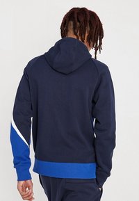 Tmavě modrá mikina s kapucí a kontrastním modrým akcentem a bílým pruhem. Má klokaní kapsu a nastavitelnou kapuci se stahovací šňůrkou. Hladká textura.