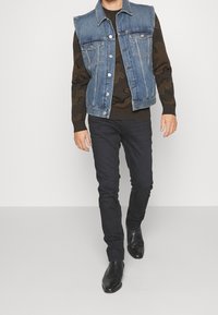Jeansweste in hellblauer Farbe mit abgenutzten Details, getragen über einem braunen Camouflage-Pullover. Schwarze enge Hosen und schwarze ankle boots.