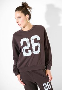 Felpa marrone con una grafica bianca "26" consumata sul davanti, dotata di scollo rotondo e polsini a costine. Indossata con pantaloni abbinati.