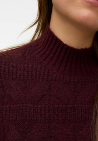 Gruby sweter wykonany na drutach w kolorze bordowym, z wysokim prążkowanym kołnierzem i złożonym, teksturowanym wzorem na całej powierzchni. Miękki, wełnopodobny materiał.