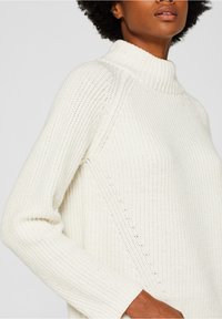 Pull en maille de couleur crème avec un col montant, une texture côtelée et des manches longues. Présente des détails de couture diagonale pour un design supplémentaire.