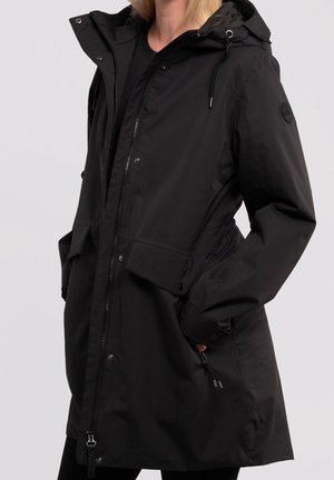 Vrouw draagt een zwarte regenjas met capuchon, voorzien van een ritssluiting aan de voorkant, drukknoopsluitingen, grote klepzakken en verstelbare trekkoordjes aan de mouwen en capuchon.