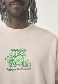 Beige sweatshirt featuring a green graphic design with a playful character and objects, including a soccer ball and a bottle.