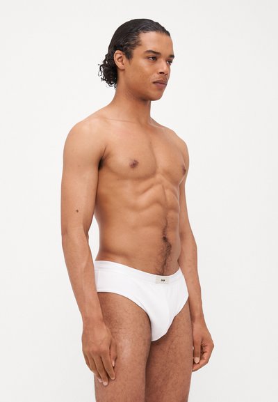 Caleçons blancs avec une texture lisse et une coupe ajustée, présentant une ceinture subtilement marquée, modélisés par une silhouette masculine de profil.