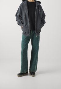 Felpa grigia oversize con tasche frontali, abbinata a jeans in denim verde a gamba larga e scarpe nere con suola color beige su uno sfondo neutro.