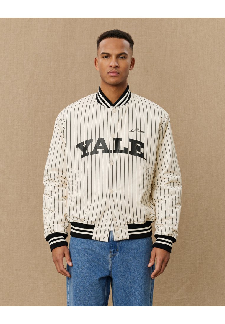 Chaqueta bomber de rayas crema con un cuello y puños ribeteados en negro, con la inscripción "YALE" impresa de forma prominente en el frente.