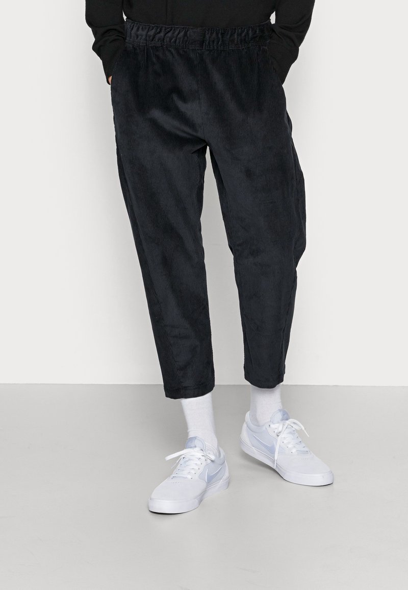 Tmavě námořnické kordové kalhoty se stahovacími pasem, tapered nohavicemi a jemným vertikálním žebrováním. Doplněné o bílé tenisky.