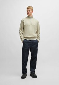 Felpa zip-up beige chiaro con zip a un quarto e una patch testurizzata sul petto, abbinata a pantaloni cargo in denim scuro.