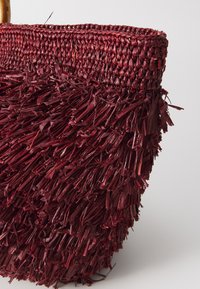 Piros szőtt táska fonott anyagból, amelynek alján texturált rojtos részlet található, és sima, kerek teteje van.