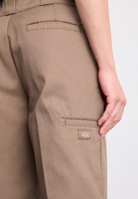 Pantaloni cargo in cotone marrone con tasca laterale e patch con logo. Texture liscia con vestibilità rilassata e dettagli di design funzionali.