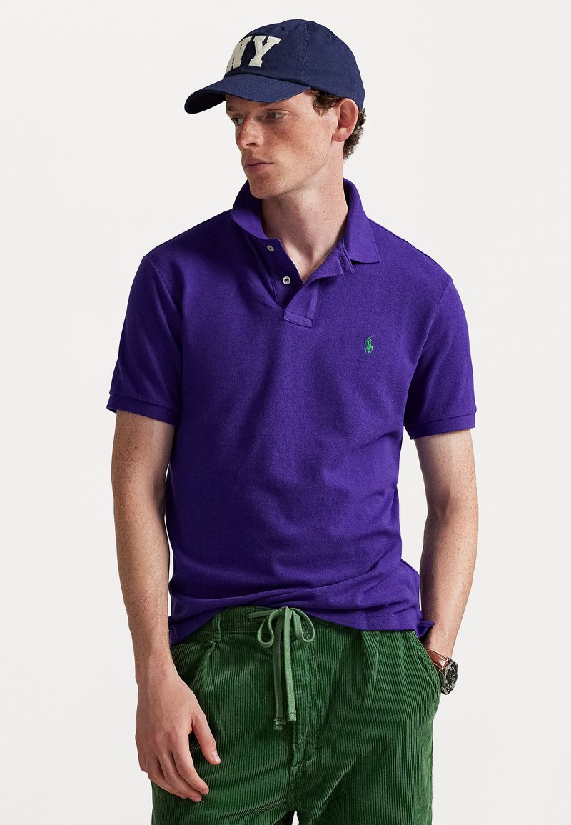 T-shirt polo roxo com mangas curtas e gola, apresentando um logótipo verde. Combinado com calças de veludo cotelê verde texturizadas e um boné azul-marinho.