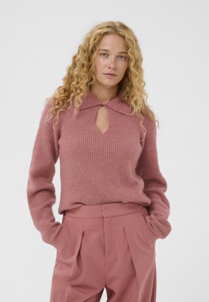 Donna con capelli biondi ricci che indossa un maglione a coste rosa polveroso con colletto e apertura a chiave, e pantaloni rosa a vita alta plissettati.