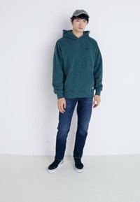 Sudadera color agua con un bolsillo tipo canguro y un logo, combinada con jeans azules y zapatillas negras. Una gorra ligera completa el atuendo.