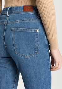 Calças de ganga azuis de cintura alta, com um patch de couro na parte de trás e um único bolso traseiro com costura branca.