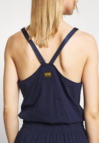 Vrouw met schouderlang blond haar, gekleed in een marineblauw mouwloos topje met gekruiste bandjes en een gele G-Star Originals-label op de achterkant.