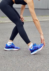 Personne en leggings noirs étirant sa jambe en touchant sa chaussure de course bleue et violette sur une surface en asphalte en plein air.