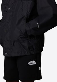 Svart vindjacka i glänsande material, med tryckknappar, elastisk nederkant och en "WINDWALL"-logotyp på ärmen. Matchad med svarta shorts.