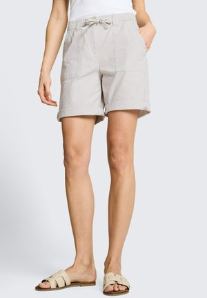 Vrouw die lichtbeige shorts met koord en opgerolde zomen draagt en beige slippers, staande tegen een effen achtergrond.