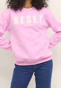 Mujer con cabello rizado que lleva una sudadera rosa con el texto "RESET RECHARGE" y jeans azules, con las manos en las caderas.