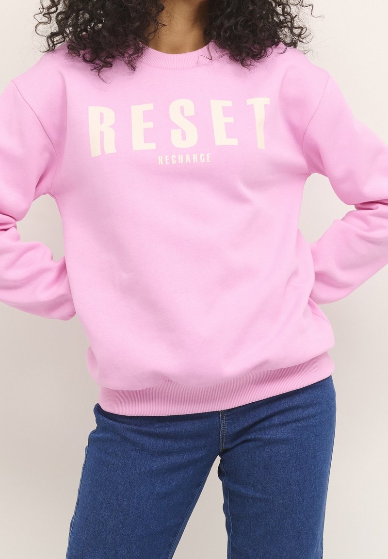 Mujer con cabello rizado que lleva una sudadera rosa con el texto "RESET RECHARGE" y jeans azules, con las manos en las caderas.