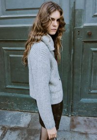 Joven mujer con cabello largo y ondulado, vestida con un suéter gris de punto y una falda negra, de pie frente a grandes puertas verdes.