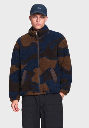 Homme portant une veste polaire bleue et marron à motifs avec une fermeture éclair à l'avant, un pantalon cargo noir et une casquette bleu marine avec un texte blanc.