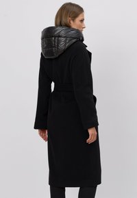 Czarny płaszcz z mieszanki wełny z puchowym czarnym nylonowym kapturem. Posiada pasek w talii, długie rękawy oraz długość do kolan. Gładka faktura.