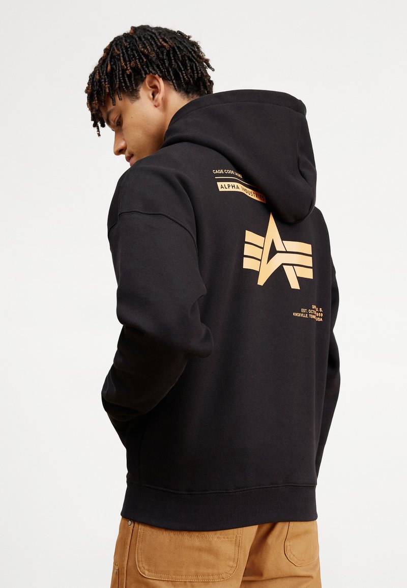 Black hoodie with a front pocket, featuring a large gold logo on the back and branding details on the upper left side. Cotton blend material.