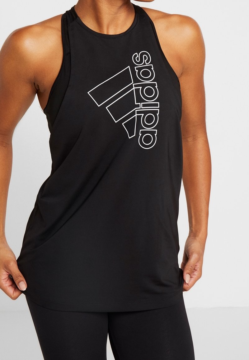 Femme portant un débardeur de sport Adidas noir avec un grand logo blanc sur la poitrine, tenant l'ourlet de la chemise.