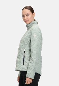 Giacca trapuntata verde chiaro con colletto alto, tasche con zip e un sottile dettaglio del logo; presenta un design elegante e aderente.