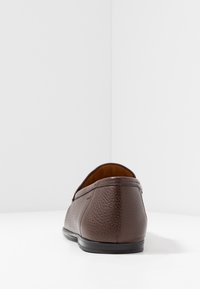Mocassins en cuir marron avec une surface texturée, un bout rond et une semelle en caoutchouc noir, vus de l'arrière sur un fond blanc.