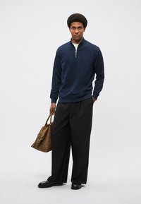 Tmavomodrý pulóver so zipsom pri krku z mäkkej látky, v kombinácii s voľnými čiernymi nohavicami. Model drží hnedú tašku s koženými detailmi. Čierne topánky dopĺňajú vzhľad.