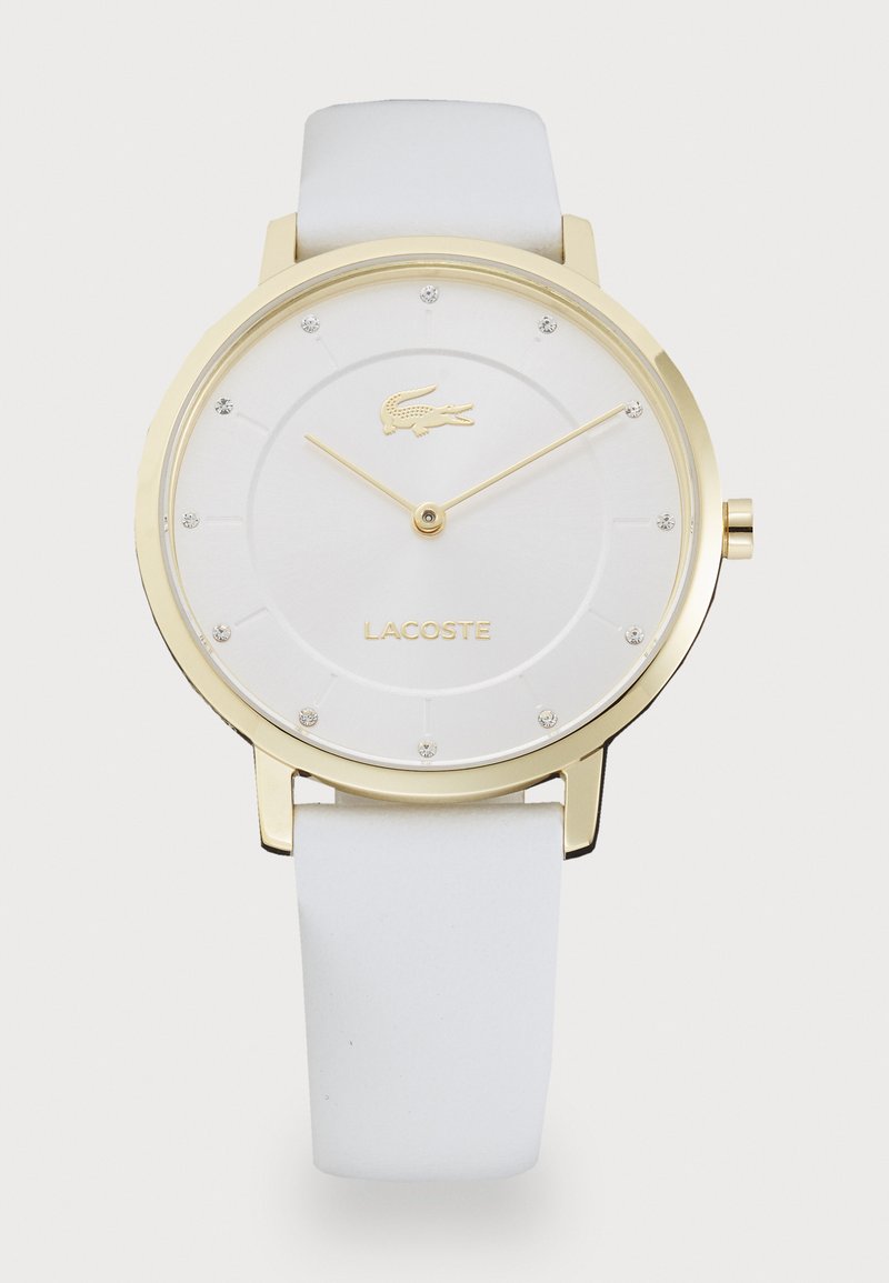 Guldtonad klocka med en vit rund urtavla, som har en Lacoste-logotyp, kristalltimmarkörer och ett vitt silikonband.