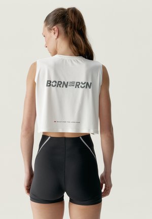 Žena s hnedými vlasmi v kučeri, nosiaca biely bezrukávový crop top s textom "BORN TO RUN" a čierne športové šortky, pohľad zo zadu.