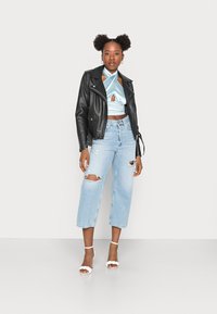 Jaqueta de cabedal preta de motociclista, blusa de cetim azul-claro com um laço à frente, calças de ganga largas de denim claro com desgastes e sandálias de tacão brancas com tiras.