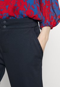 Svarta byxor med en slät textur, prydda med tv å knappdetaljer och sidofickor, som matchas med en blus med röd och blå mönstring.