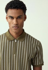 Jongen met krullend haar die een olijfgroene gestreepte polo met korte mouwen en een rits aan de voorkant draagt, kijkt recht vooruit.