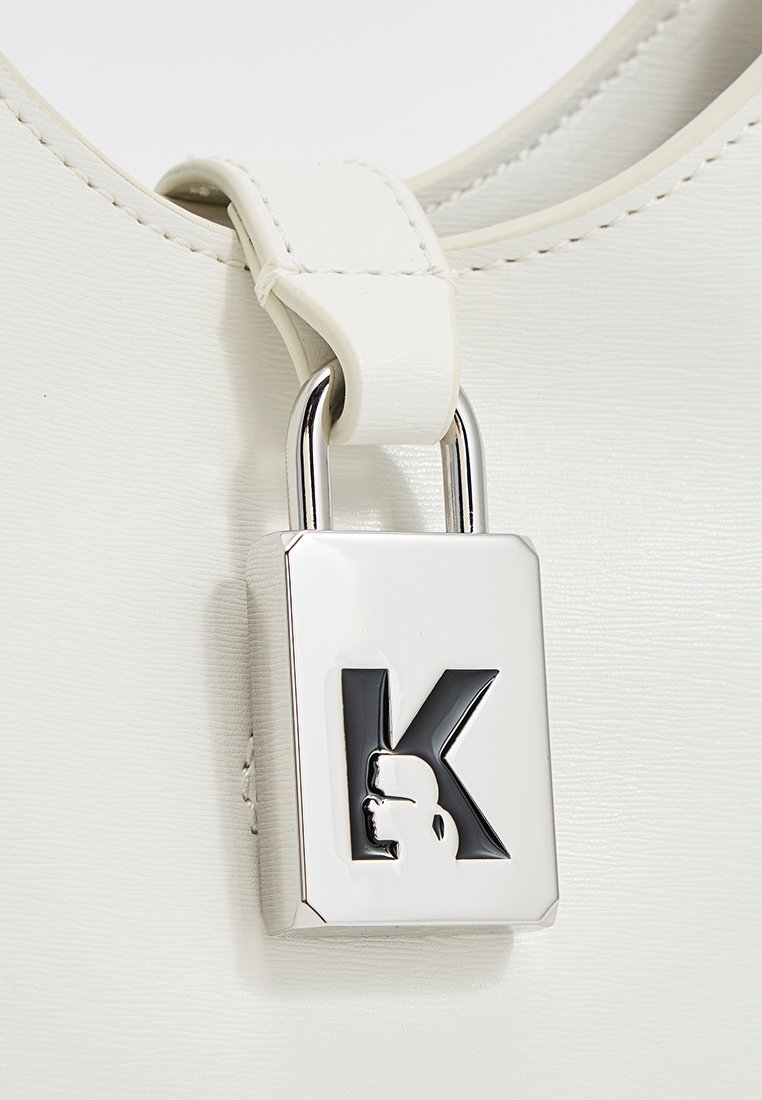 Sac à main blanc au toucher lisse, avec un accent de cadenas argenté et un logo noir. Sangle réglable attachée avec un connecteur blanc.