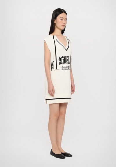 Knit dress, cream-colored with black accents, features a deep V-neck, short sleeves, and a graphic print. Worn with black flats.