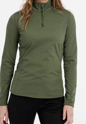 Femme portant un pull vert olive à manches longues avec une fermeture éclair sur un quart et un col haut, associé à un pantalon noir.