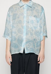 Camisa de manga corta en un patrón floral azul claro y gris, con botones en la parte delantera y un bolsillo en el pecho, confeccionada en una tela texturizada y ligera.
