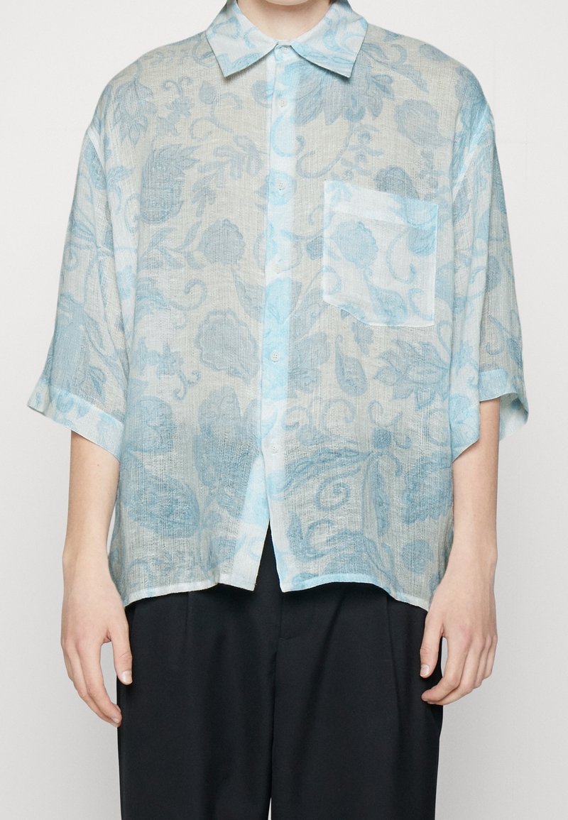 Camisa de manga corta en un patrón floral azul claro y gris, con botones en la parte delantera y un bolsillo en el pecho, confeccionada en una tela texturizada y ligera.