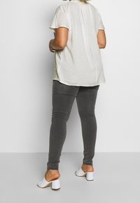 Bluzka z krótkim rękawem w kremowym kolorze z teksturowanym wzorem, zestawiona z dopasowanymi szarymi dżinsami i białymi mule z blokowym obcasem. Widoczny guzik z tyłu.