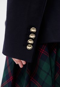 Σκούρο μπλε ύφασμα μανίκι με τέσσερα χρυσά κουμπιά, που διαθέτει περίπλοκα σχέδια. Μια πράσινη και σκούρα μπλε καρό φούστα είναι μερικώς ορατή από κάτω.